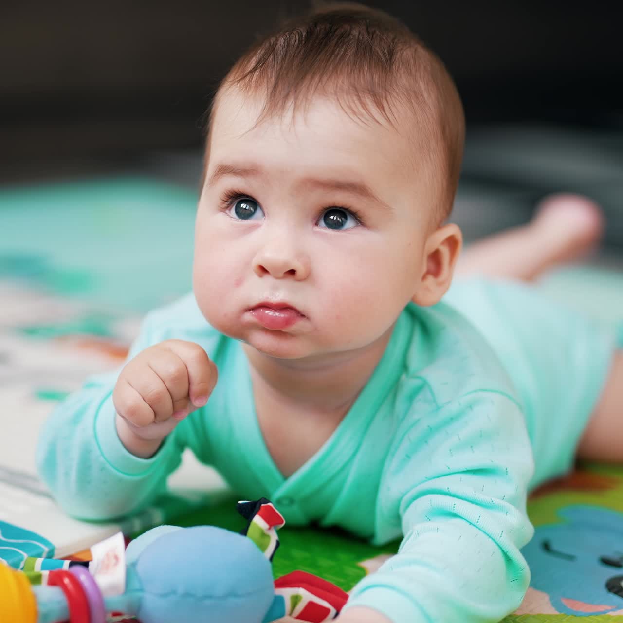 Adorable beautiful baby in blue clothes lies on the floor. Lovely kid lies among the toys on a colorful mat looking around. Close up. Blurred backdrop