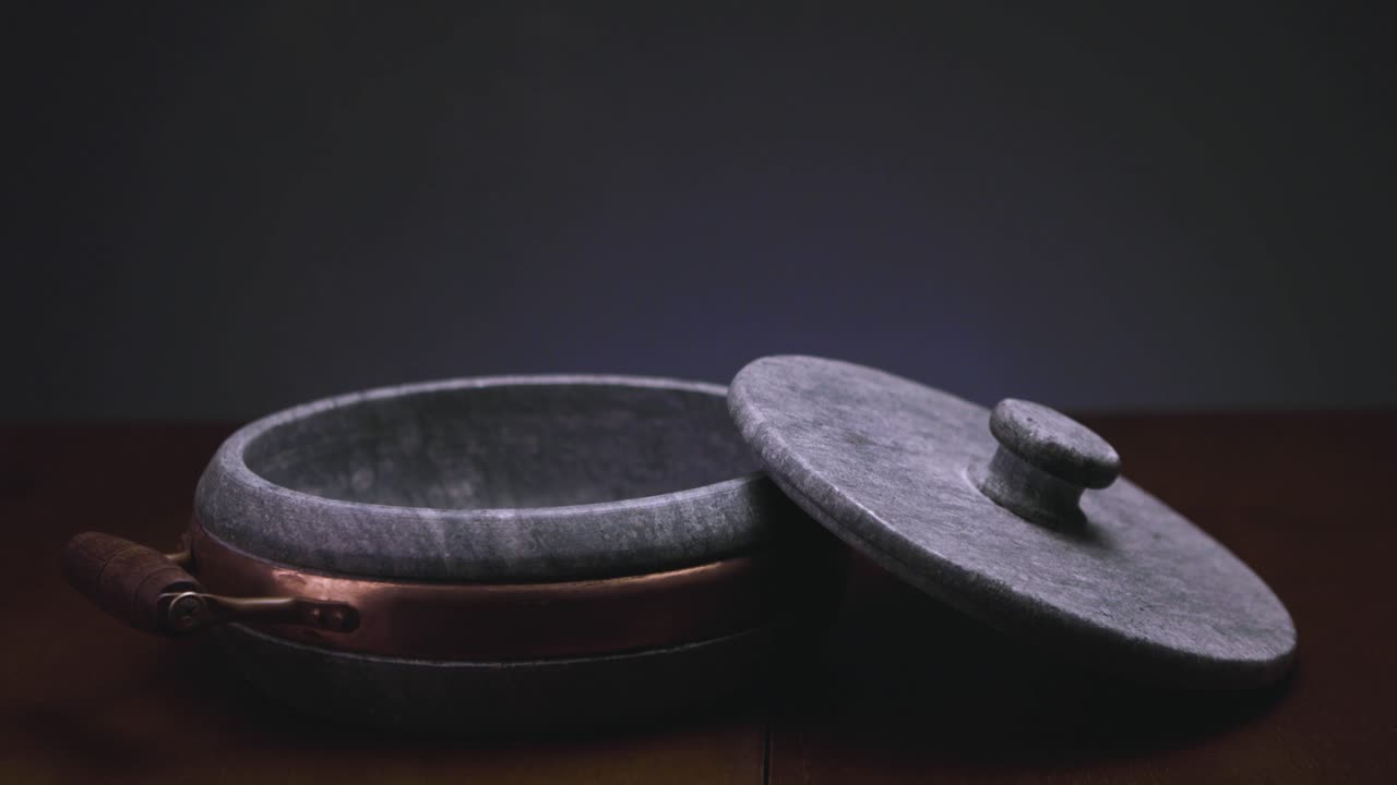 Steam coming out of a smooth round soap stone casserole with brass element and wooden handle with the lid resting half on it on a brown wooden table surface seen from above looking into the pan