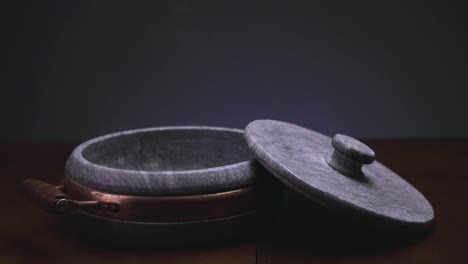 steam coming out of a smooth round soap stone casserole with brass element and wooden handle with the lid resting half on it on a brown wooden table surface seen from above looking into the pan