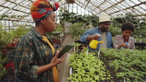 Familia-Afroamericana-Usando-Tabletas-Y-Rociando-Plantas-En-Invernadero