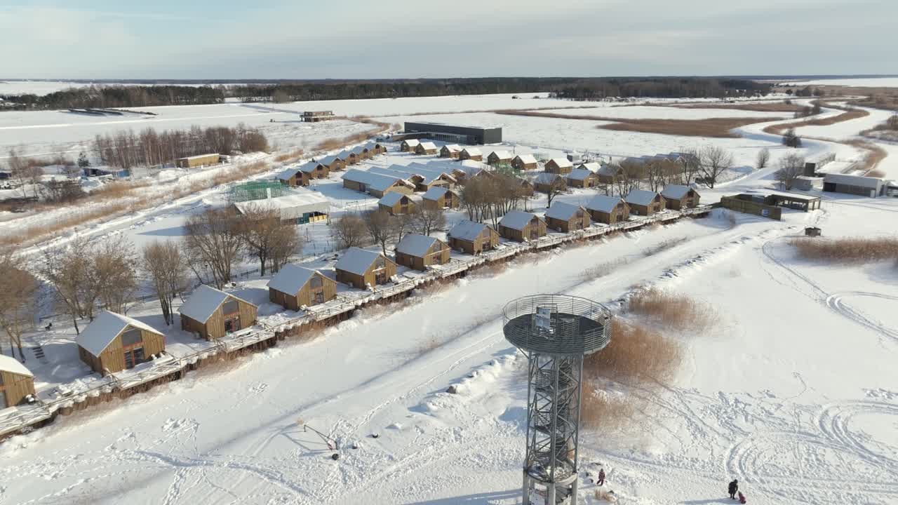 A wide drone shot revealing a cluster of wooden holiday homes nestled in a snowy landscape, featuring a modern metal observation tower and tracks leading into the white horizon