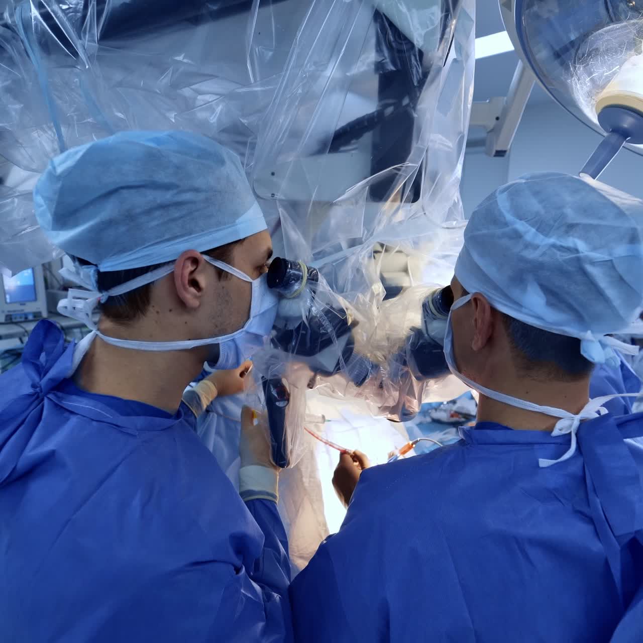 Backs of the surgeons standing at microscope looking at binoculars. Doctors move the tools slowly operating the patient