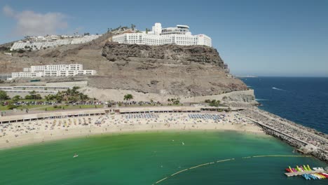 Vista-Aérea-Circular-De-La-Playa-De-Los-Amadores-En-Puerto-Rico,-Gran-Canaria-Con-Hoteles-Y-Gente-Bañándose-Al-Fondo-Al-Mediodía