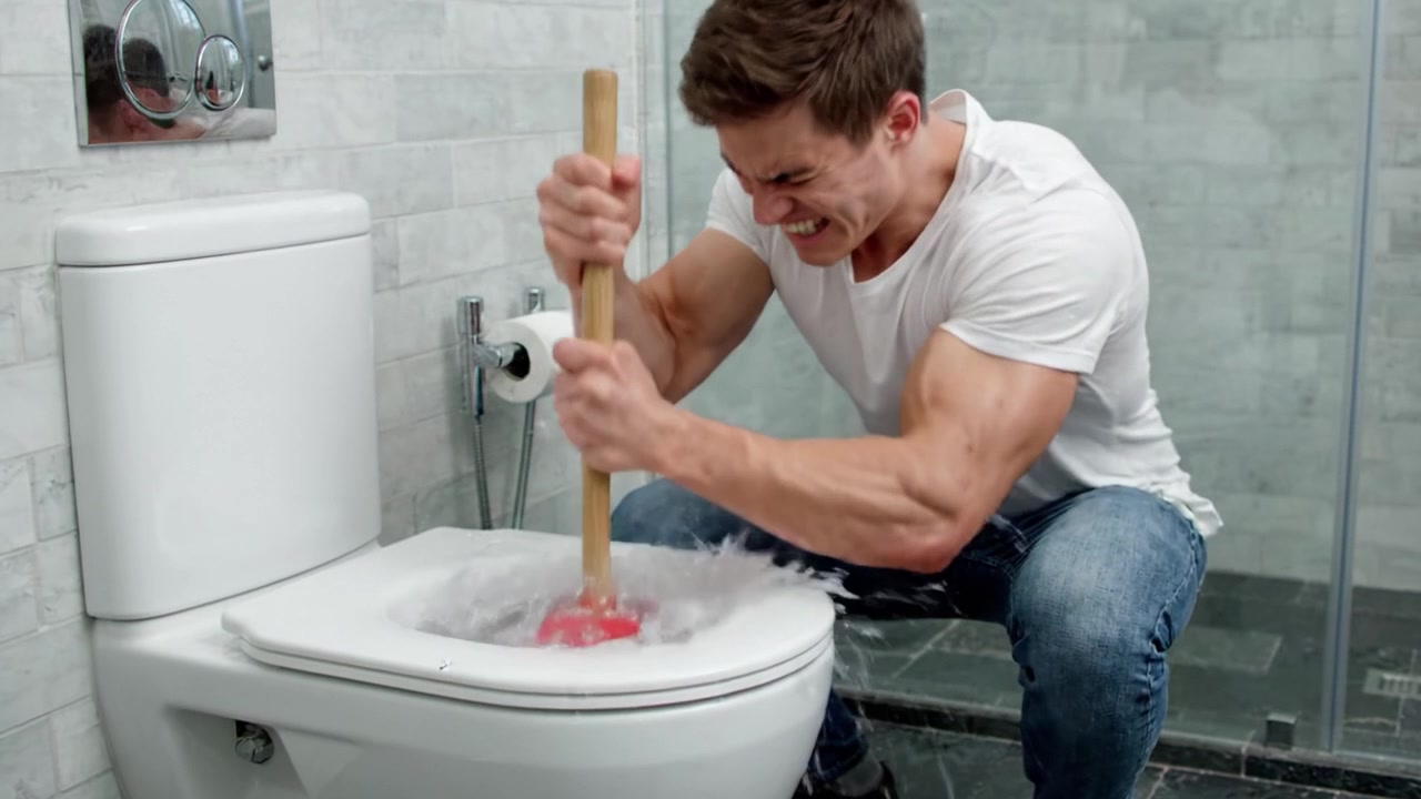 A Determined Young Man Struggles to Fix a Clogged Toilet with a Plunger, Showcasing His Physical Effort and Concentration in a Modern Bathroom Setting