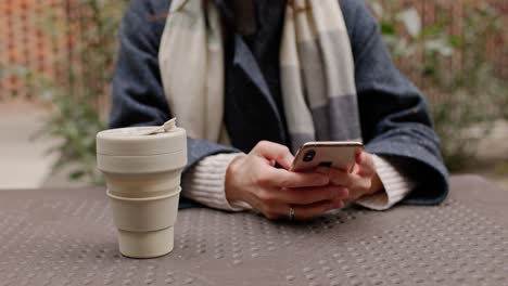 woman using phone at outdoor cafe with reusable cup