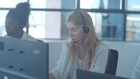 focused fair haired operator in headset taking call and talking with her colleague