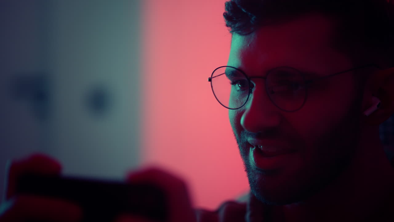 jugador alegre jugando videojuegos por teléfono inteligente en la aplicación retrato emocional de un joven guapo con gafas teléfono móvil y auriculares en el oído adicción al juego ciberdeportivo en línea ganar y alegría