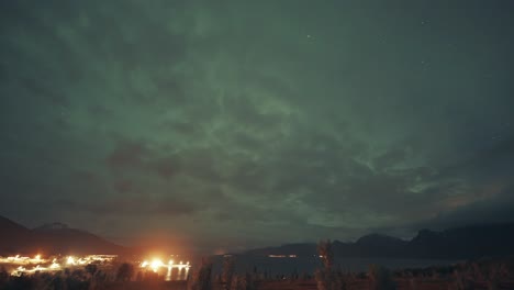 luces del norte en el cielo nublado sobre una ciudad y un puerto brillantemente iluminados en el norte de noruega