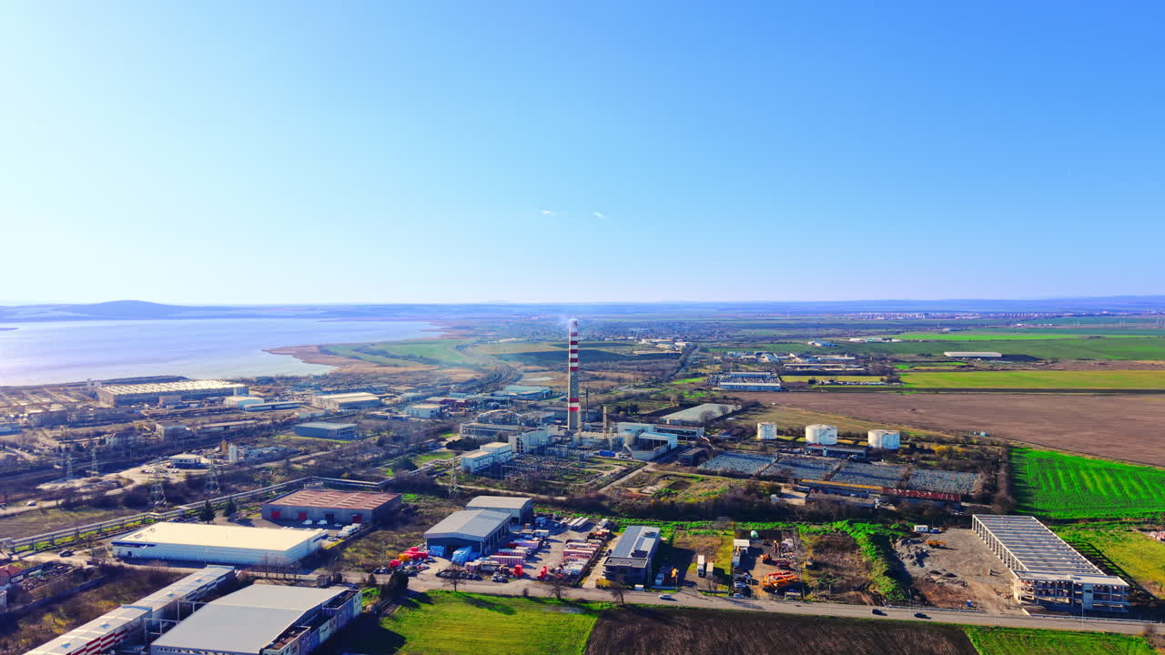 Panoramisk luftfoto av et industriområde med en høy skorstein, lagerbygninger og jordbruksområder. Industridistrikt og kraftverk nær sjøen.