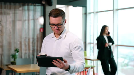 continuing work during the coffee break. man is focused on i-pad holding a paper cup. woman stand with cup speaking on the phone at blurred backdrop.