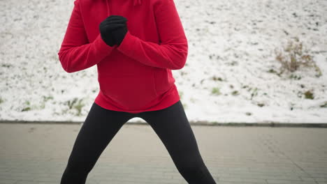 coach in red hoodie demonstrates side lunge exercise during outdoor winter workout on snow-covered paved ground surrounded by snowy hill with iron railing, in cold weather