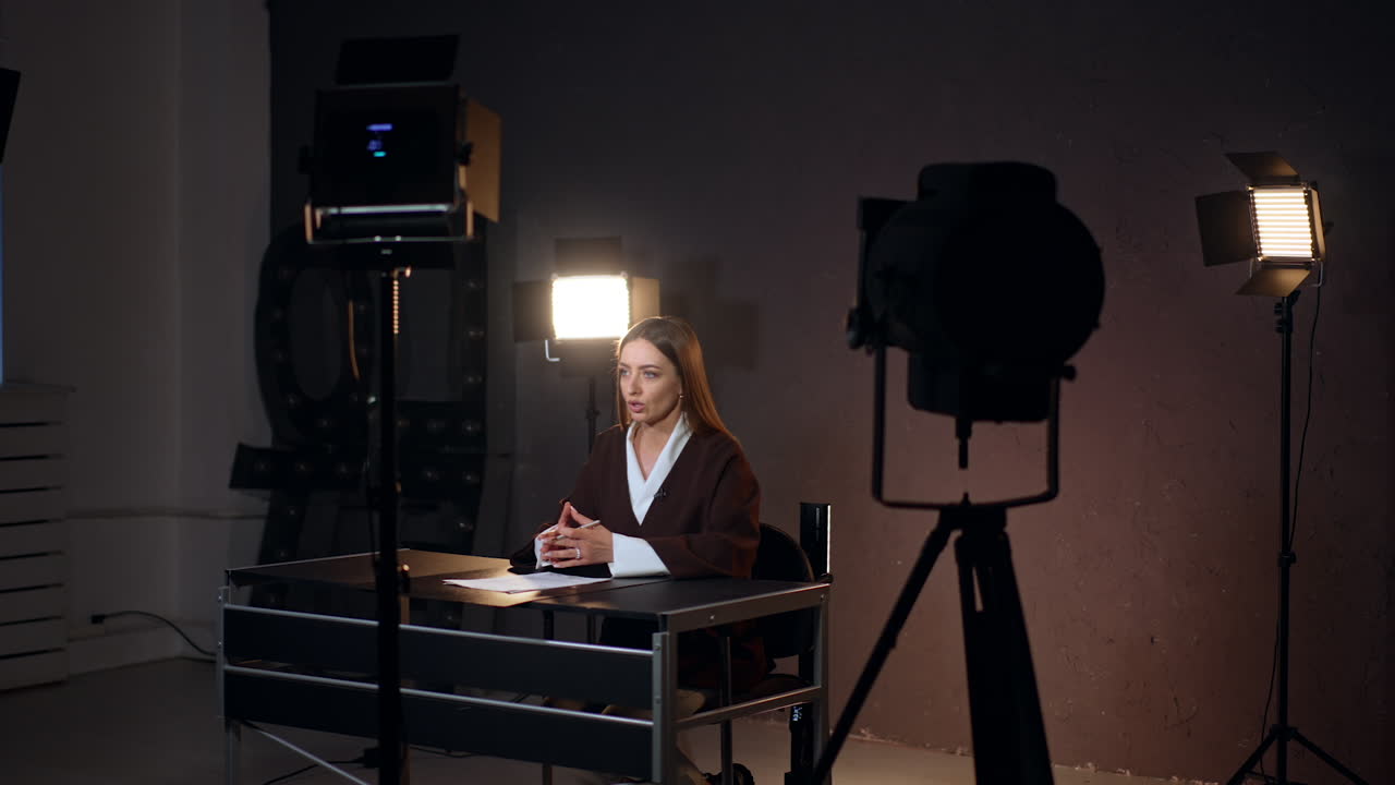 Brunette long-haired Caucasian woman sits at desk talking to camera. Footage in photo studio with soffits switched on.