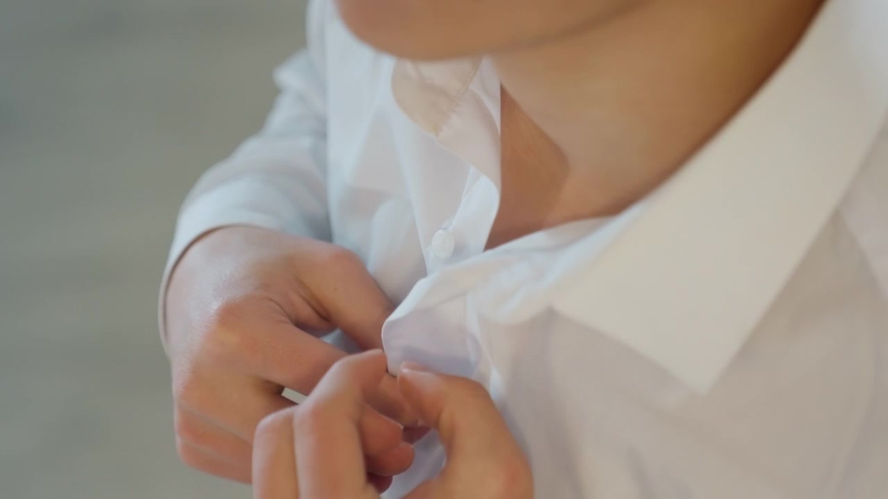 Uomo asiatico che allaccia una camicia bianca in una camera da letto luminosa, primo piano delle dita che abbottonano, sottile energia nervosa, preparazione per un colloquio di lavoro, look casual-formale, luce naturale dalla finestra, atmosfera intima e spontanea.