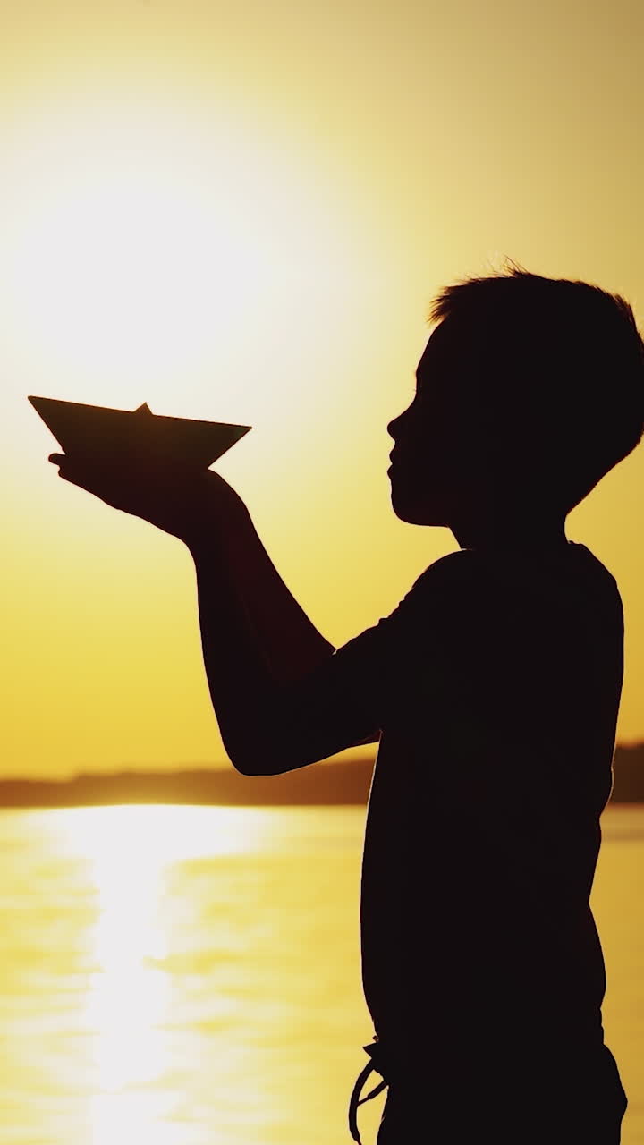 A little boy is holding an origami in the form of a paper boat in his hands and raising up it to the level of the sun on the background of the lake. Silhouette of a guy at sunset Vertical video