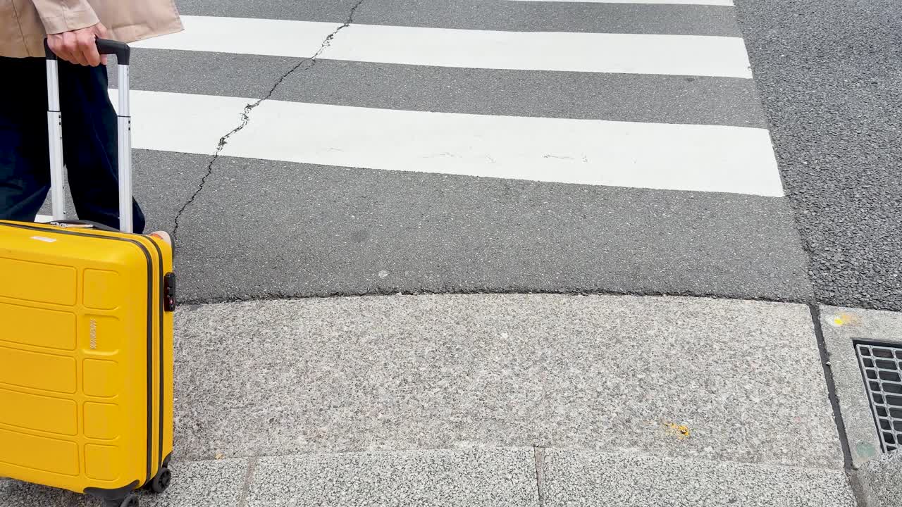 Low angle shot of a traveler pulling yellow luggage across a city street crosswalk