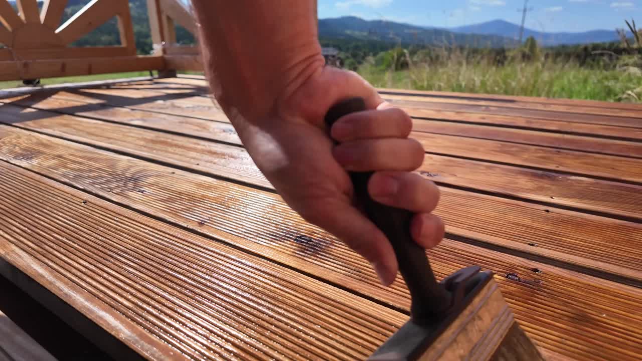 Hand and brush oils varnishing impregnating wooden terrace boards. Decorative railing, serene sky, clouds, and mountain horizon evoke home improvement and outdoor maintenance themes
