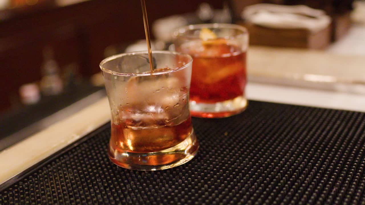 Bartender pours and stirs whiskey cocktails with ice in dimly lit, upscale bar setting
