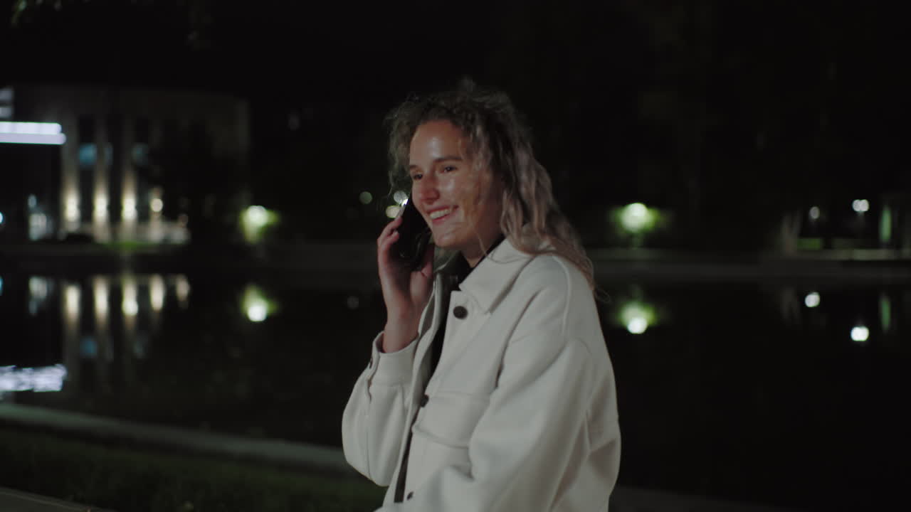 Young girl laughs while talking on phone at night with reflection near water, wearing white coat, capturing friendly warmth in softly illuminated environment, quiet ambiance, relaxed vibe
