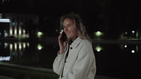 young girl laughs while talking on phone at night with reflection near water, wearing white coat, capturing friendly warmth in softly illuminated environment, quiet ambiance, relaxed vibe