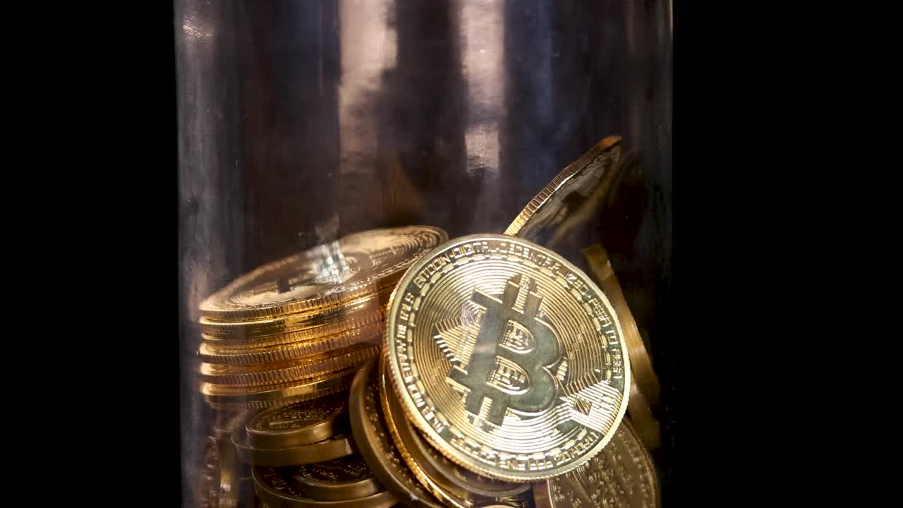 Gold-colored cryptocurrency coins fall into a glass jar, illuminated by warm, reflective studio lighting