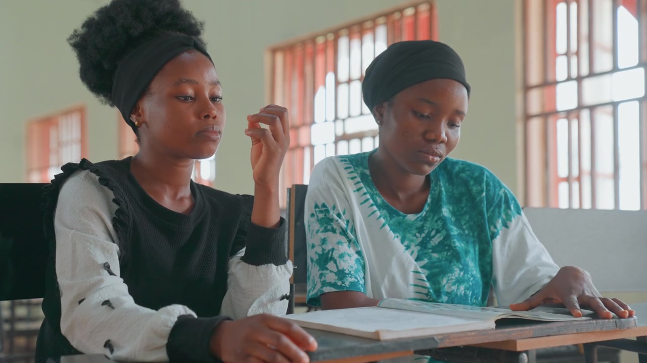 Black mother and daughter studying together at sunlit classroom desk, mother tutoring teenage child, reviewing notes and assignments, patient guidance, focused expressions, window light, cultural