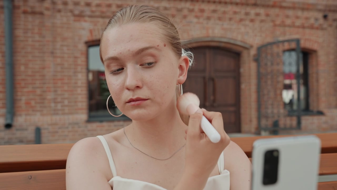 Giovane donna si trucca usando un pennello mentre tiene in mano uno smartphone, in un ambiente esterno con edifici in mattoni e panchine di legno. La donna indossa orecchini a cerchio e ha i capelli parzialmente raccolti.