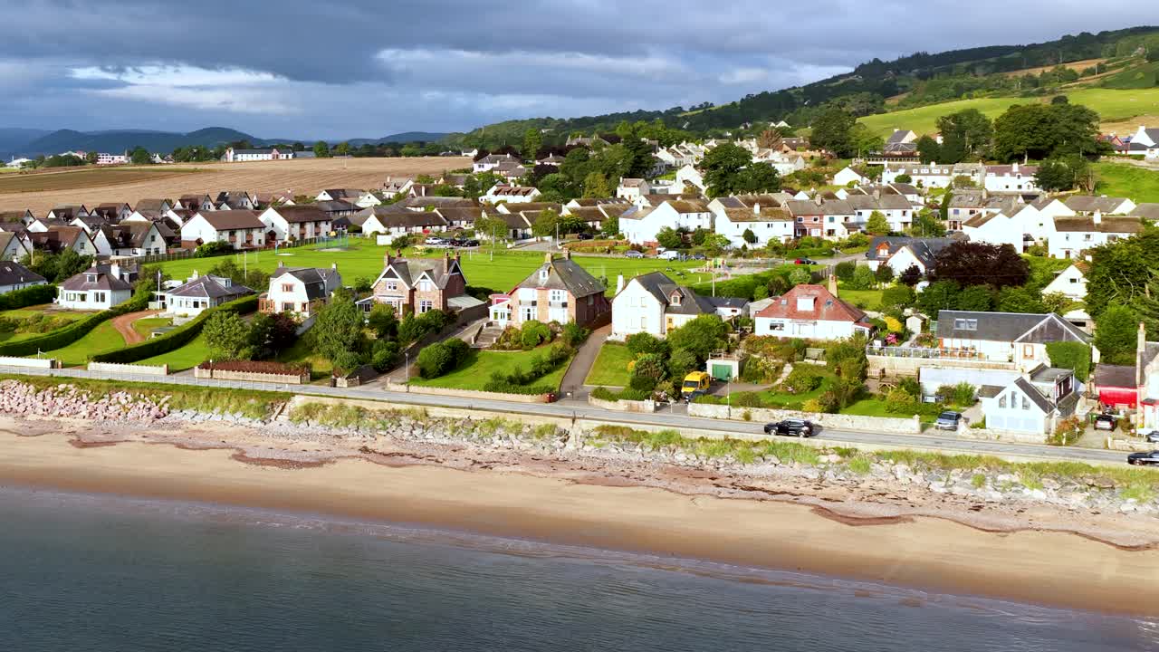 Drone glides above coastal village, revealing beachfront houses, green lawns, sandy shore, and hills
