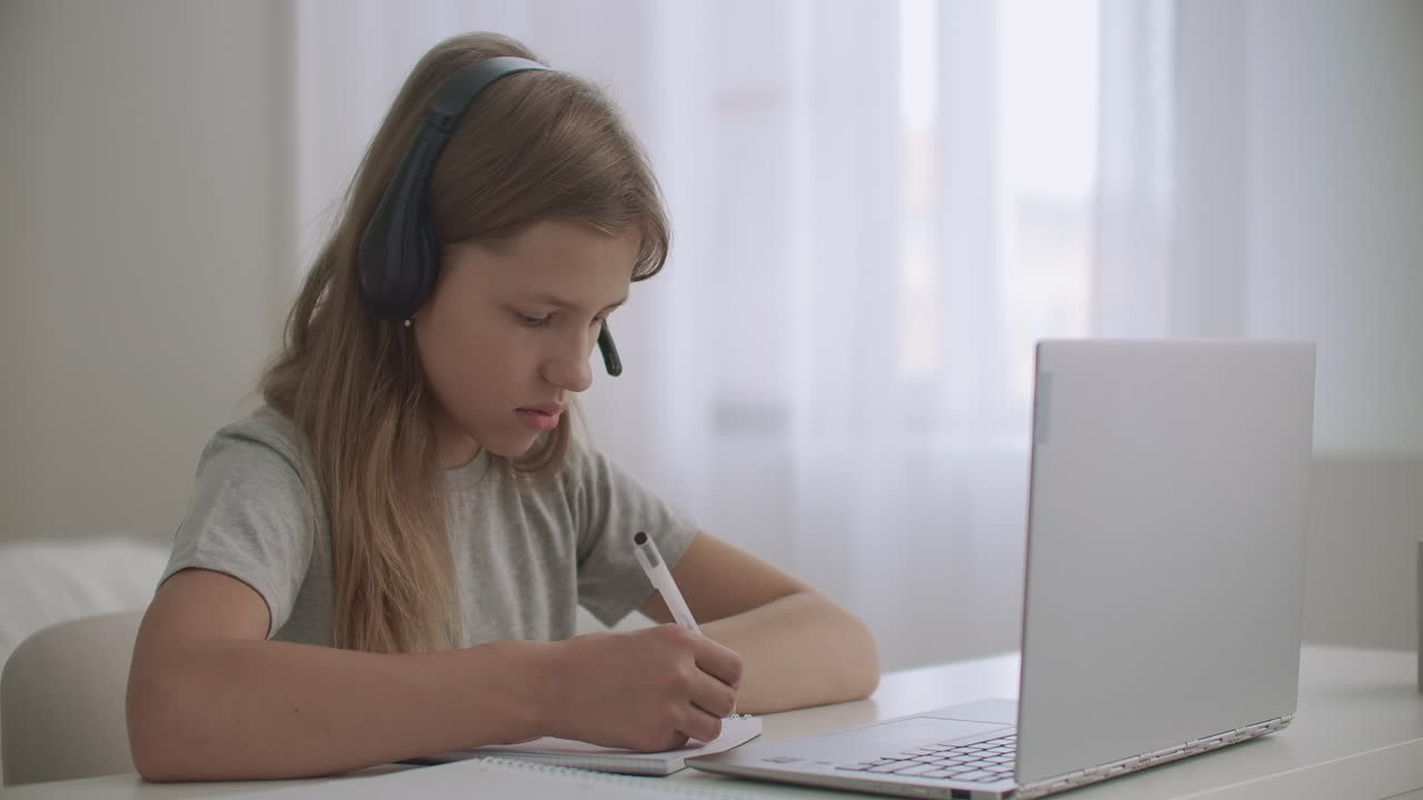 la niña está aprendiendo en línea por internet escuchando al maestro a través de auriculares y mirando la pantalla del cuaderno escribiendo notas en el libro de ejercicios
