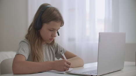 little girl is learning online by internet listening teacher through headphones and looking at screen of notebook writing notes in exercise book