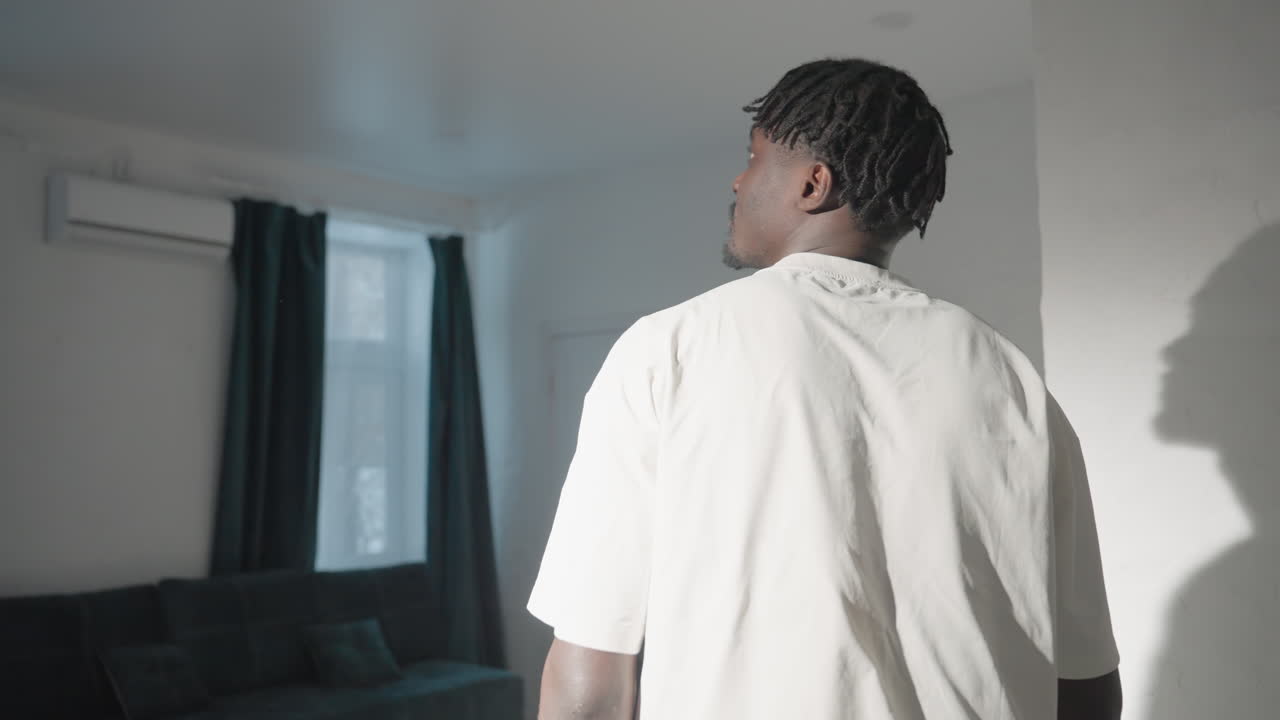 Person with short dreadlocks turning away, wearing a white t-shirt, in a modern minimalist living room. A sleek dining table, TV, and large windows with dark curtains are visible in the background