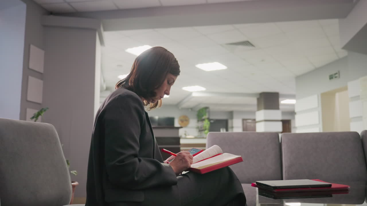 Businesswoman reading notebook in office lounge area. Female professional focused on work or study while sitting on sofa with modern office design in background. Calm and quiet workspace atmosphere