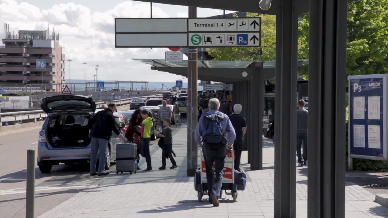 People walking with luggage at a busy airport drop-off area on a sunny day