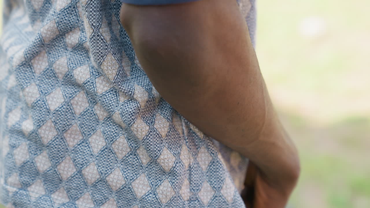 Atleta masculino negro, manos a la cintura ajustándose pantalones cortos negros, camisa estampada y fondo de parque, gesto de calentamiento previo al juego, dedos revisando el agarre y el cordón, escena de entrenamiento casual con entrenador o compañero de equipo