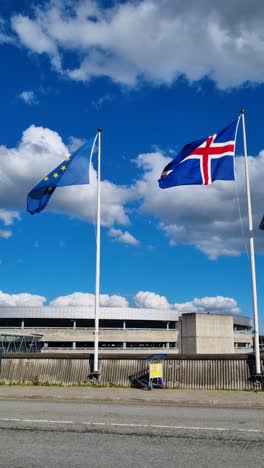 vertical shot of the icelandic and european union flags gently swaying in the wind against a partly cloudy sky, filmed at arlanda airport, sweden