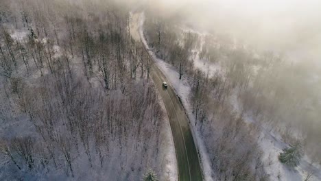 aerial view of car driving on winter country road in snowy forest covered with golden fog lit up by sun revealing the mountain in the distance