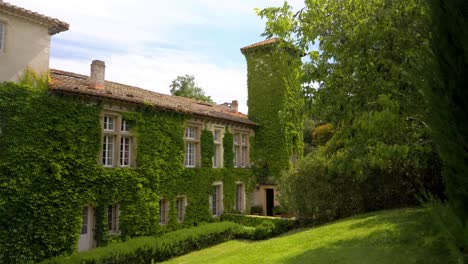 side-shot-behind-a-tree-that-reveals-a-large-house-covered-with-ivy-with-a-large-tower,-the-weather-is-sunny-and-the-wind-moves-the-leaves