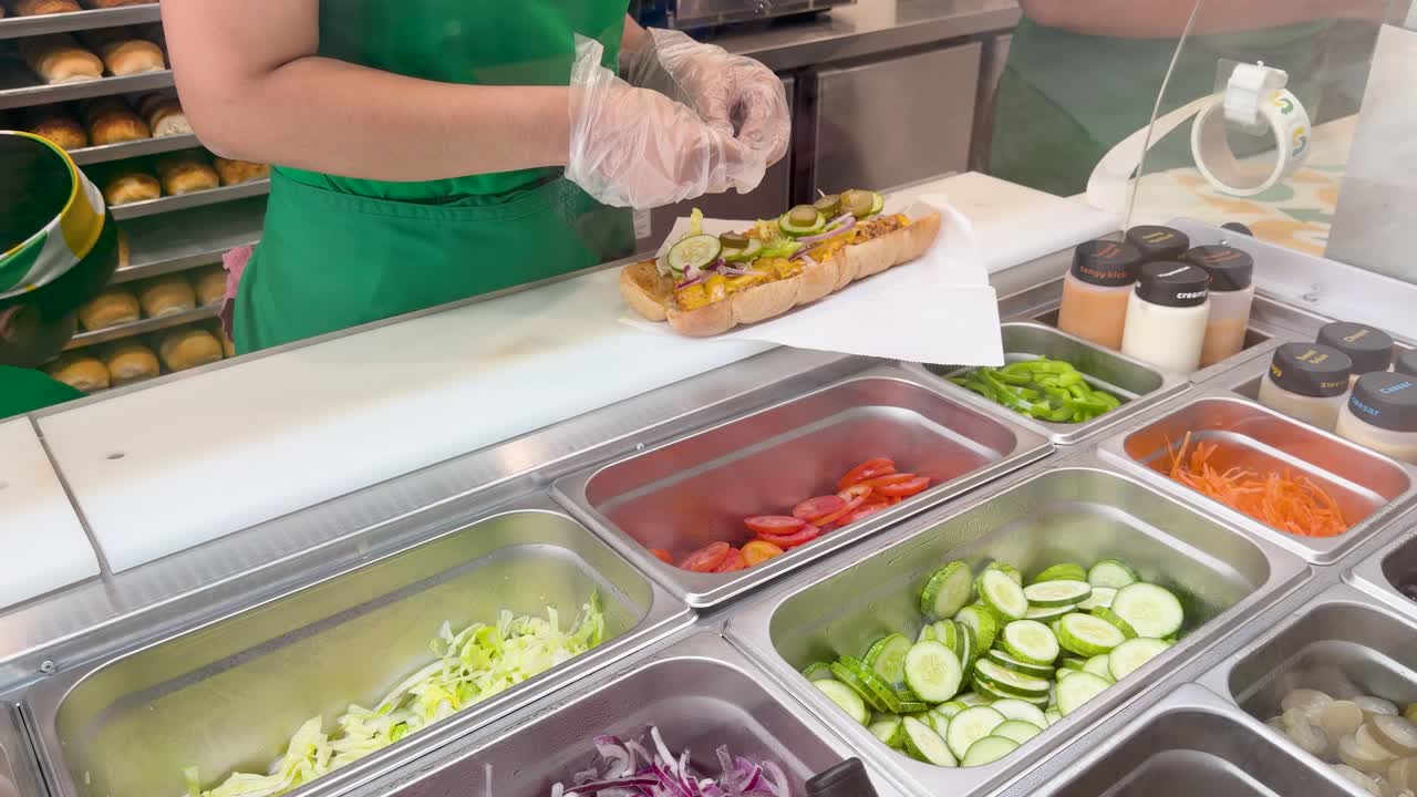 Un addetto in divisa verde prepara un panino farcito con verdure fresche dietro un bancone