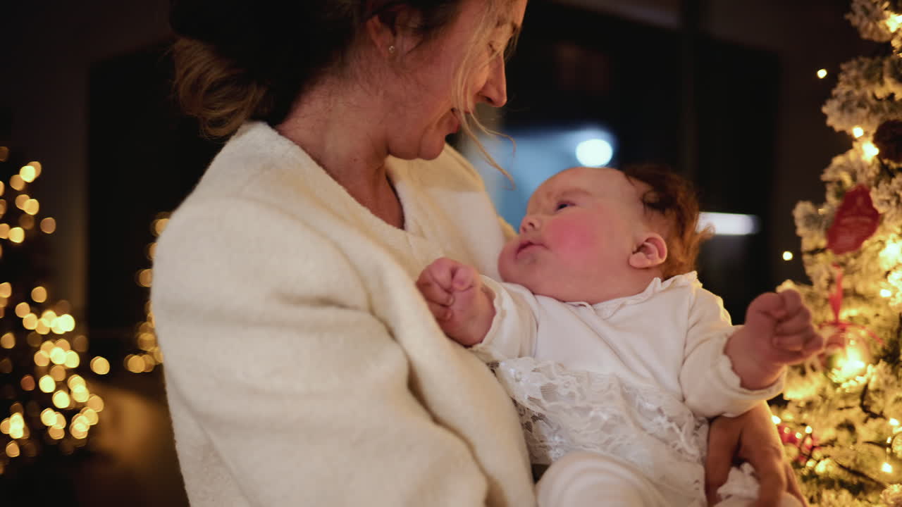 Mamma felice che tiene in braccio un bambino in casa accanto a un albero di Natale decorato con illuminazione calda e un bokeh di sfondo delicato che crea un'atmosfera intima e festosa