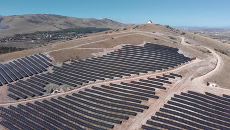 Orbit-Drone-over-solar-power-park-farm-row-panels-church-on-hills-sunny-day