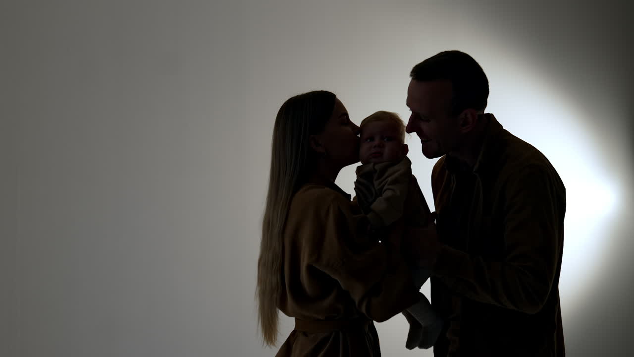 Mother and father hold their little baby. Parents kiss their son on two cheeks. Silhouette of happy family.