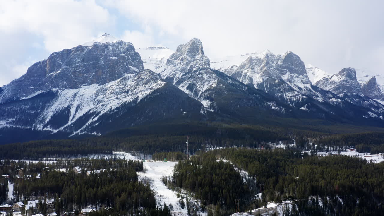A drone flies left across the snowy Alberta landscape, showcasing jagged mountain peaks, dense forests, and winter cabins nestled in the valley.