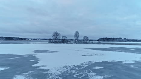 Drone-flying-towards-frozen-islet,-thin-snow-layer-over-icy-lake