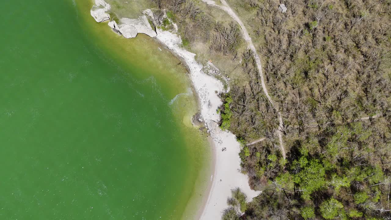 Drone yläpuolelta seuraa Steep Rockin rantaa Manitobassa, näyttäen kalkkikivikallioita, Manitobajärven vihreää vettä, puiden läpi kulkevia polkuja ja rantaviivaa kesäpäivänä.