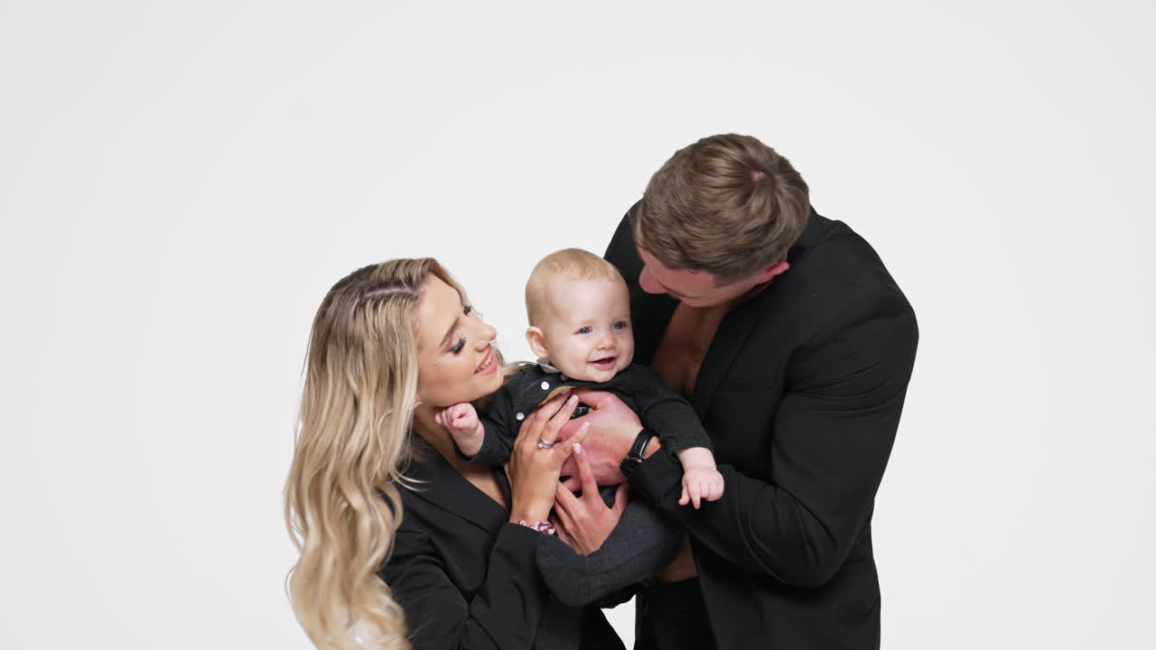 Beautiful Caucasian couple with their little baby. Loving parents kiss their adorable smiling infant son. White backdrop.