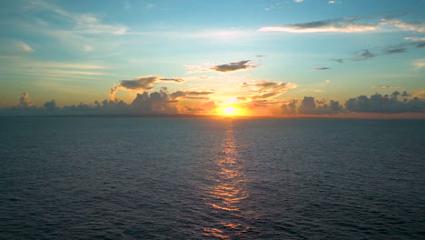 Spectacular-view-of-a-sunset-on-the-high-seas,-beyond-an-ocean-horizon-in-the-Caribbean,-blue-sky-above,-the-clouds-resembling-a-city-skyline,-sunlight-reflecting-on-the-waves,-scenic-static-FHD-shot