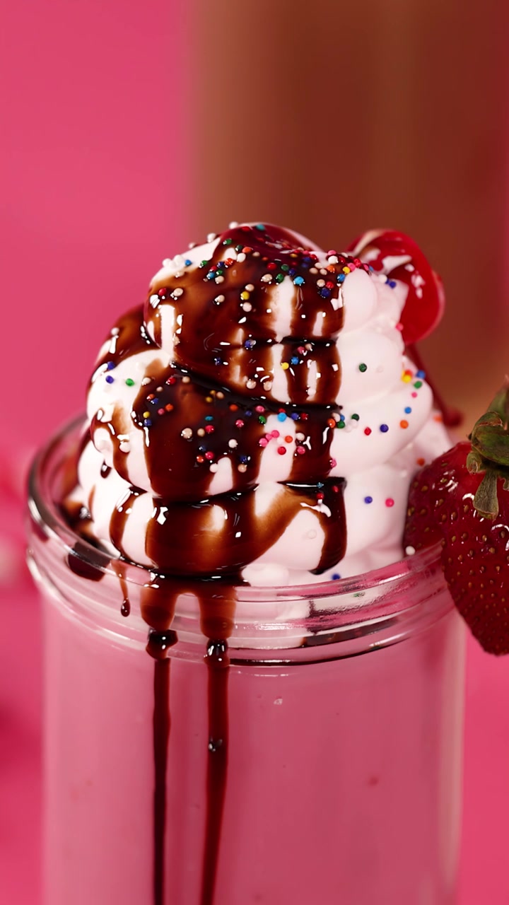 Primo piano di un milkshake cremoso alla fragola con panna montata, sciroppo al cioccolato, ciliegia, codette, sfondo rosa.
