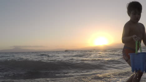 niño pequeño agarra un balde de agua de mar, con puesta de sol en el fondo en kalamata, grecia