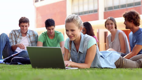 blonde student using laptop with classmates sitting behind on grass