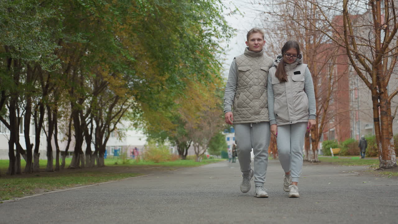 Young lovers walking side by side through serene park path, holding hands and enjoying peaceful stroll together in matching casual outfits, surrounded by trees, greenery, and urban buildings