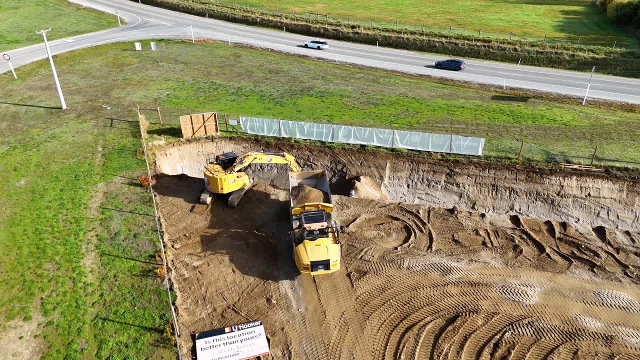 Aerial footage of an excavator and dump truck working on a construction site under bright daylight in Cromwell, New Zealand